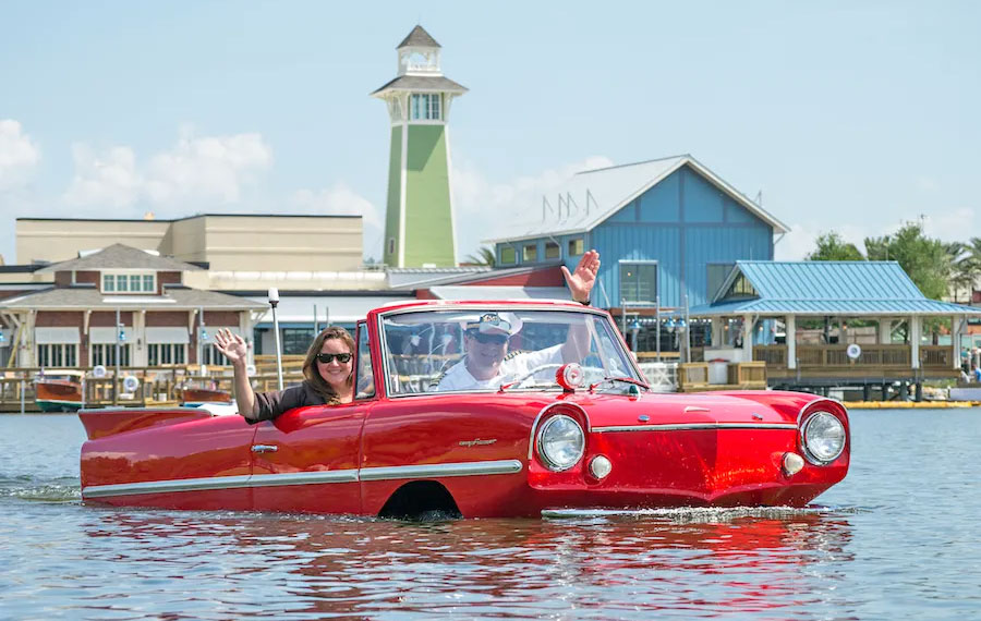 couple driving amphibious red car boat waving with buildings in background at boathouse disney springs