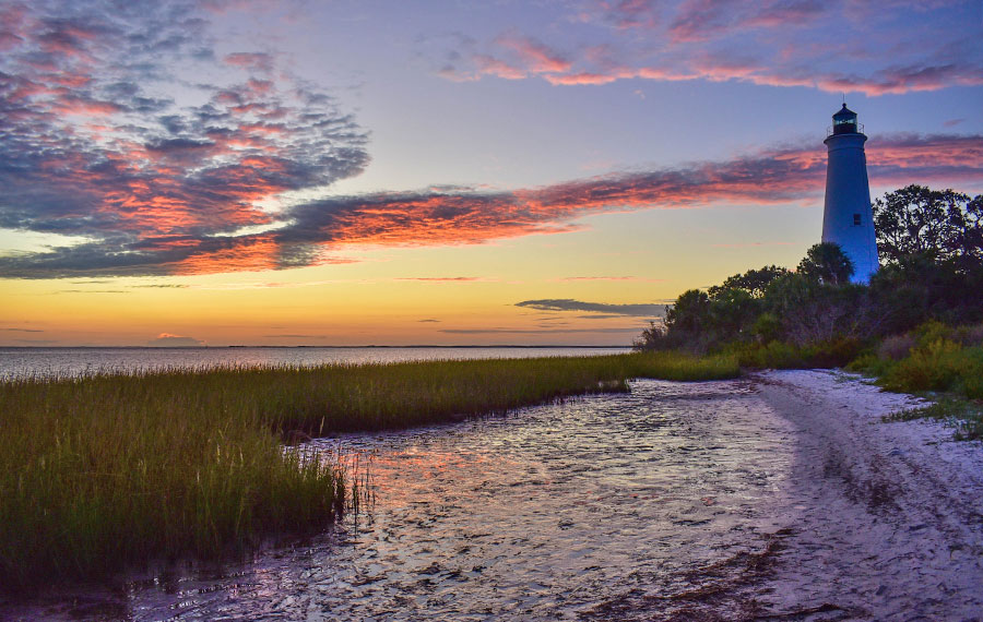 dimly lit lighthouse on right side of beach with grass and gulf under colorful sunset sky with streaked clouds st marks lighthouse near tallahassee