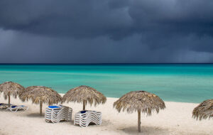 view of sandy beach with tiki shelters and loungers stacked near turquoise water with dark storm clouds offshore