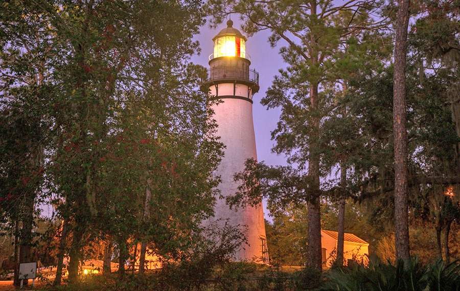 uphill view of lighthouse lit up amid treeline with purple twilight sky at amelia island lighthouse