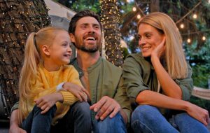 parents with young daughter laughing while sitting in night camp area with string lights on trees