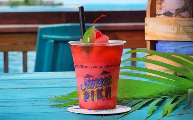 red cocktail drink in branded cup with straw on blue table at sunset pier key west