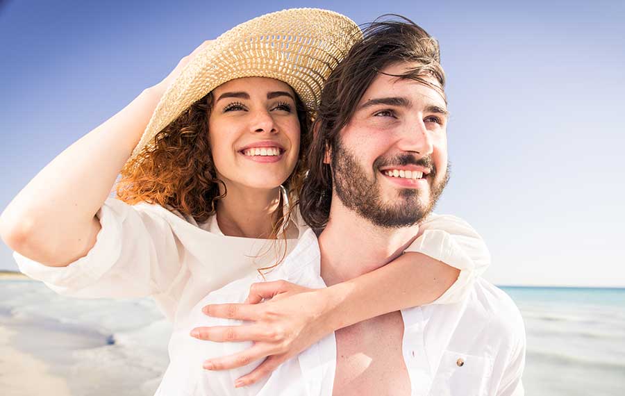 smiling couple with woman in straw hat piggybacking on breezy beach